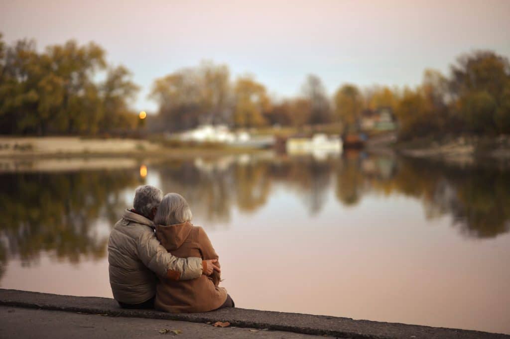 An elderly couple sits by a calm lake at dusk, embracing as they watch the peaceful water.