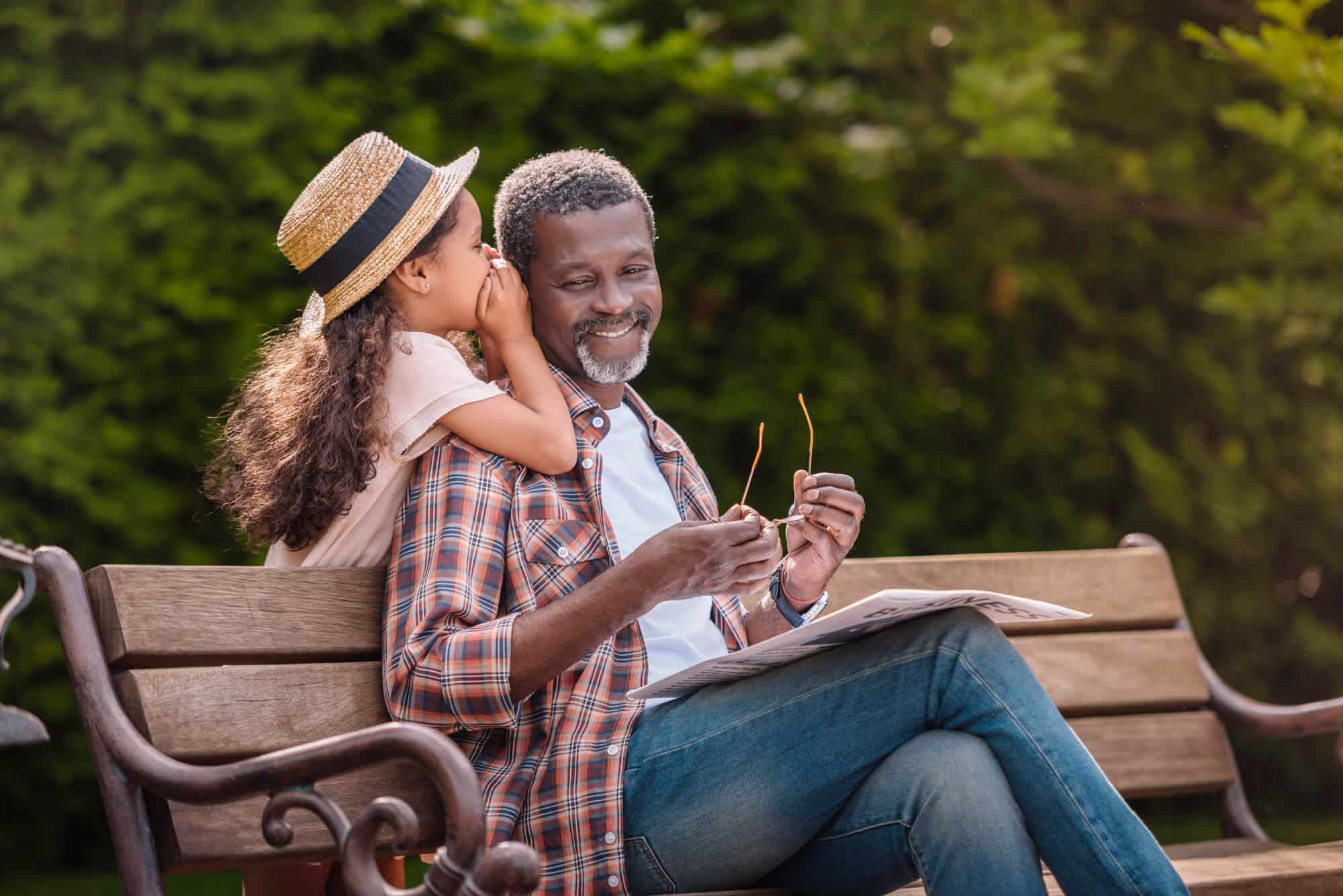 A young girl whispers into her grandfather’s ear as he smiles, sitting on a park bench with a newspaper in hand in Boise, ID