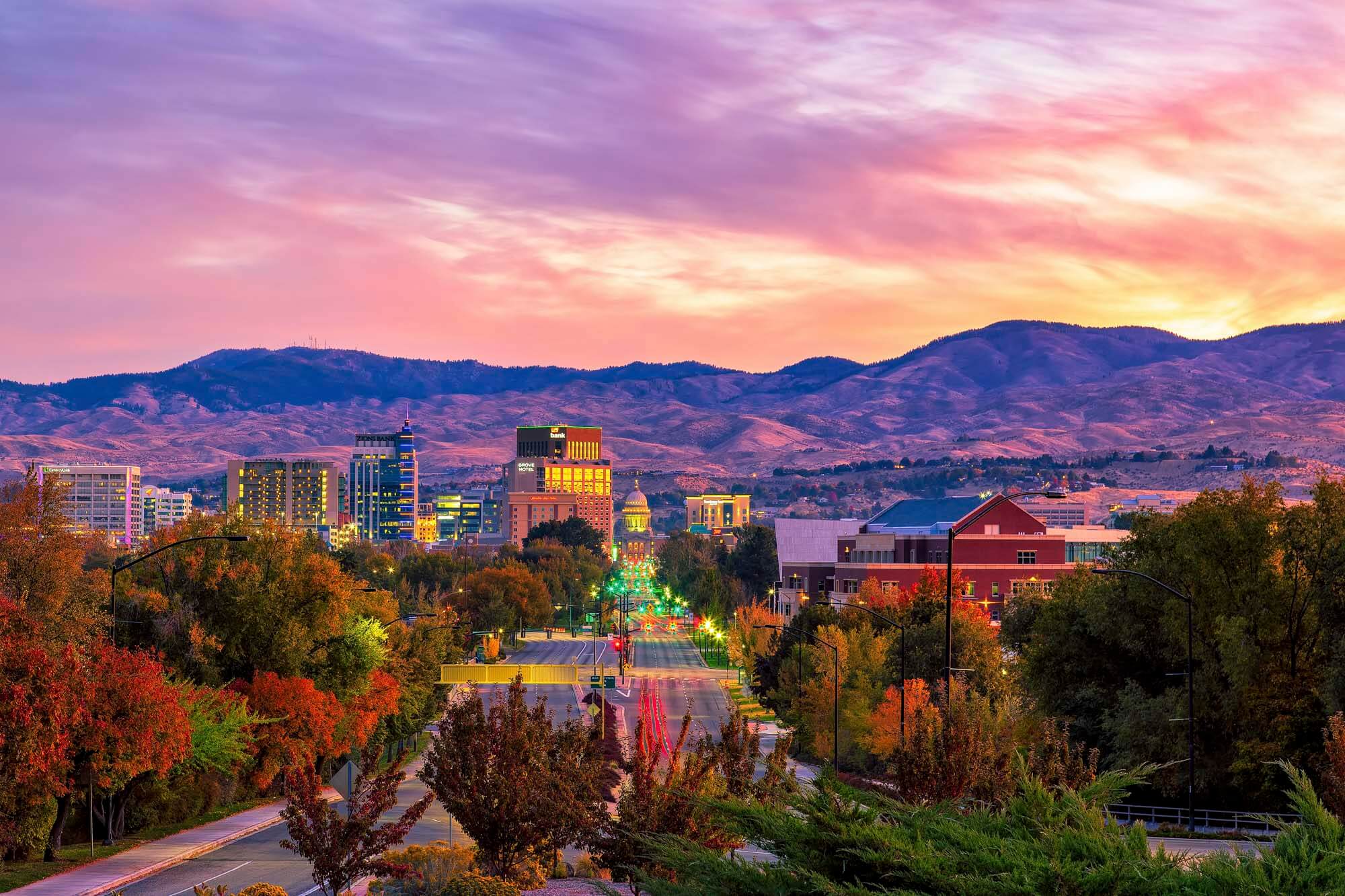 A vibrant cityscape at sunset with illuminated buildings, tree-lined streets, and mountains in the background.
