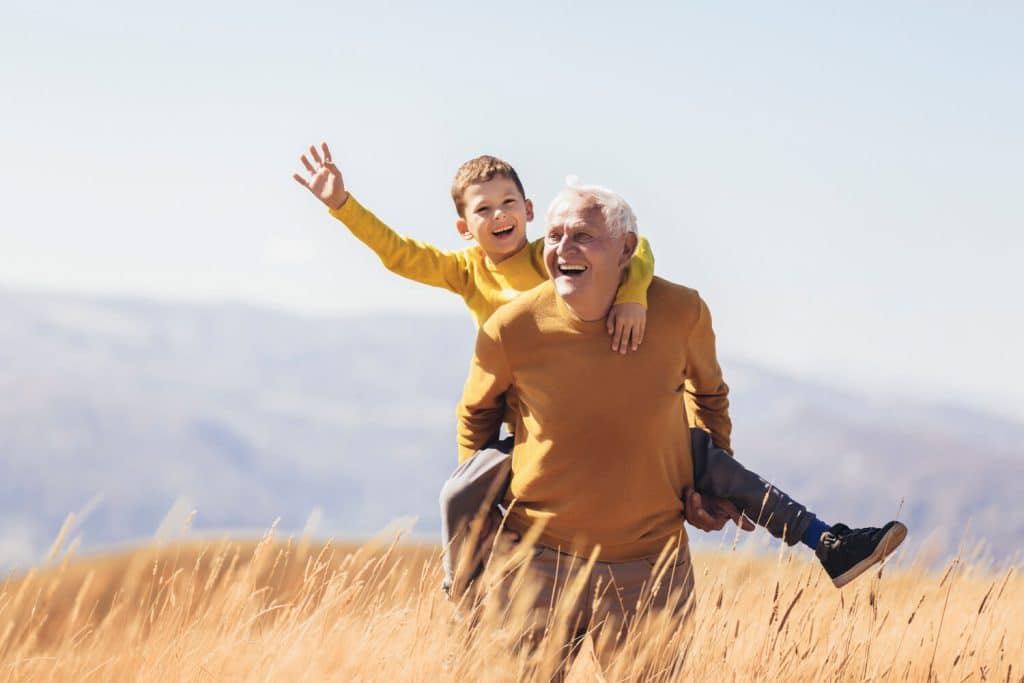 A grandfather carries his smiling grandson on his back through a golden field, both wearing matching yellow sweaters.