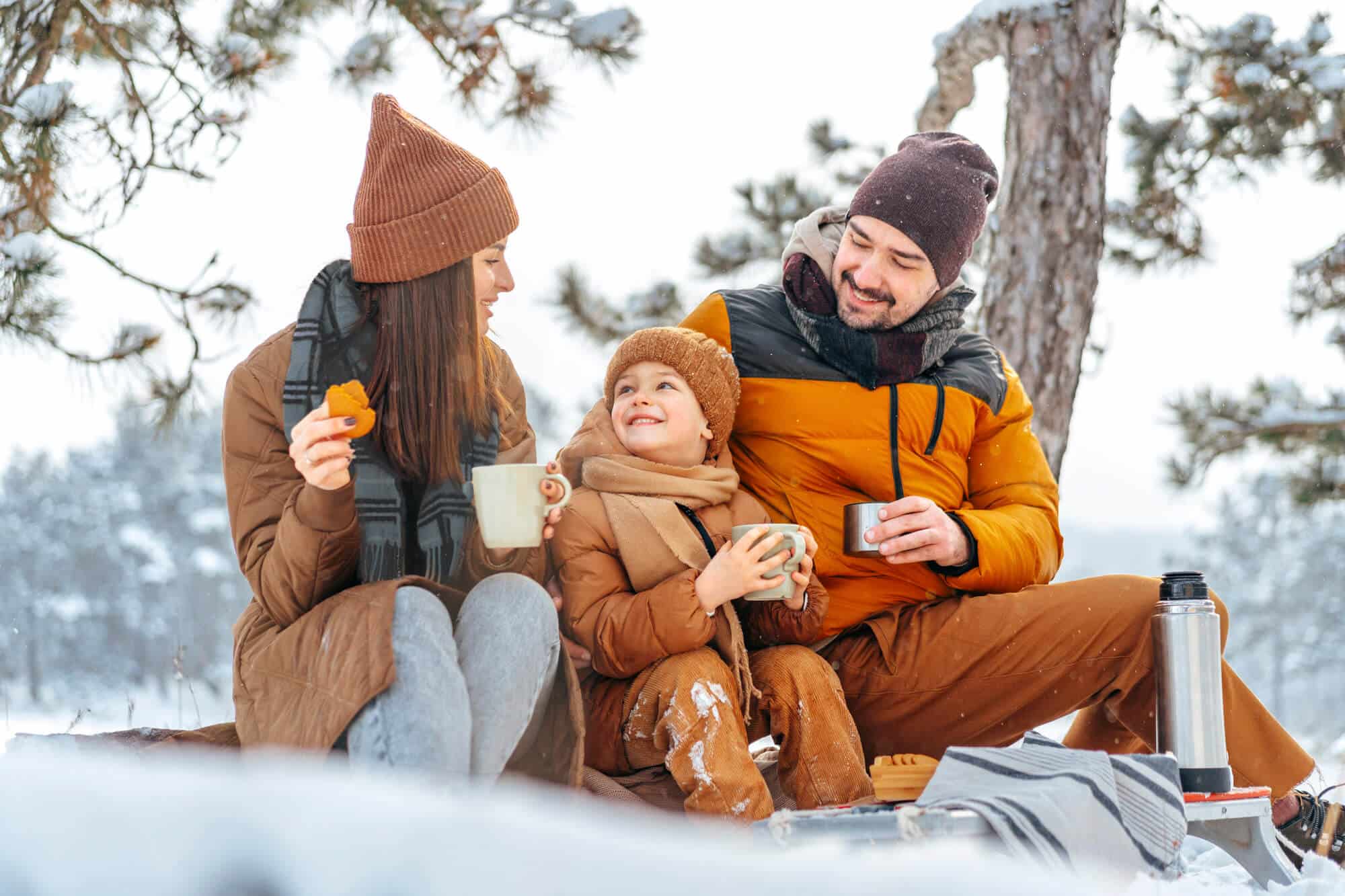 A family enjoys a winter picnic outdoors, dressed warmly, drinking hot beverages, and smiling at each other.